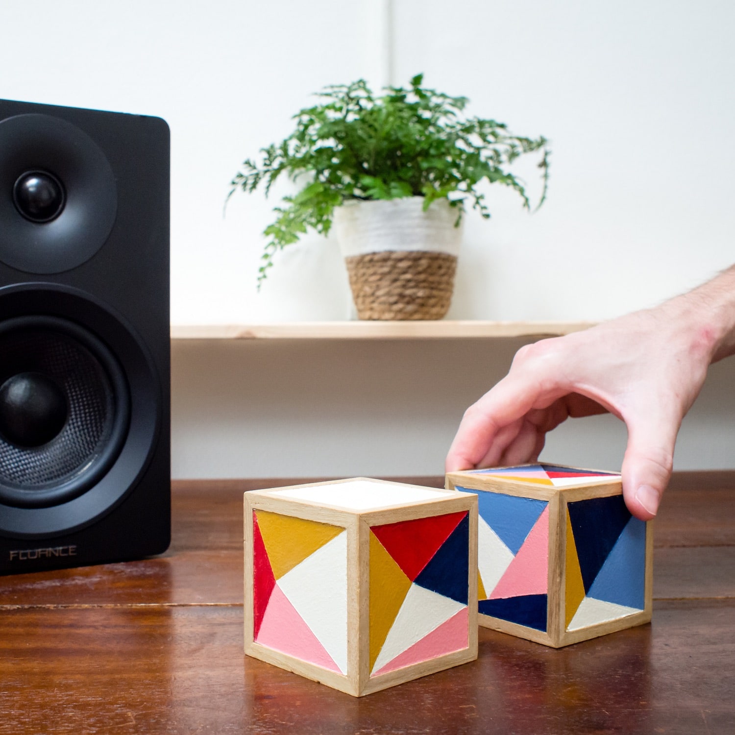 A photo of a hand grabbing ml.cubes prototype on a wooden desk next to a music speaker and fern plant.