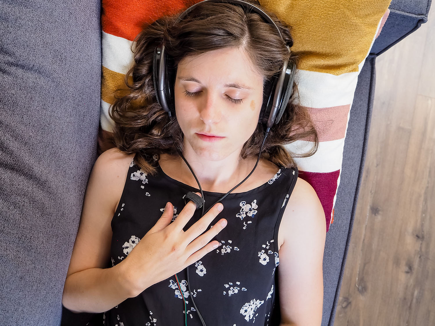 A photo of a woman in a flower dress with over-ear headphones on listening to her pulse from a sensor which is on her finger.