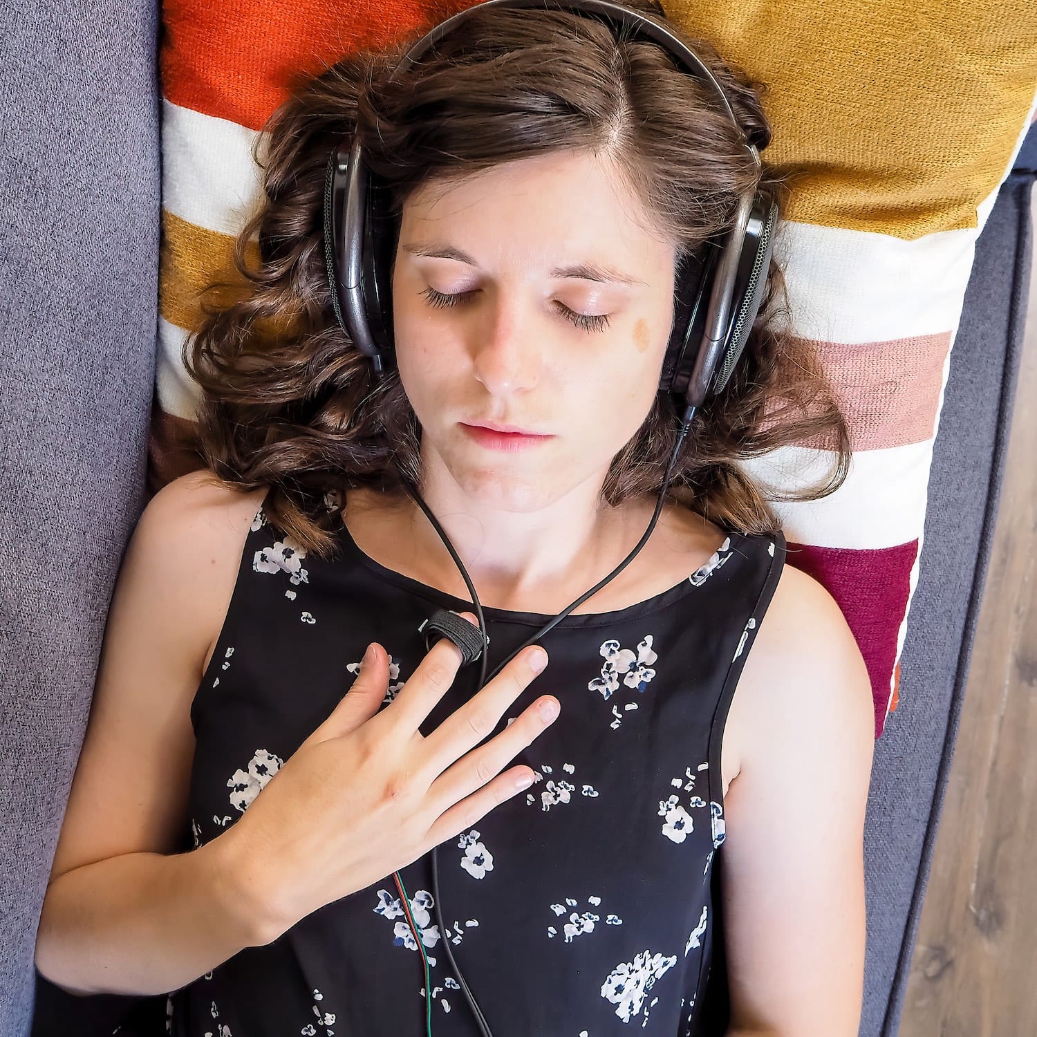 A photo of a woman in dress wearing large headphones and fingertip pulse sensor laying with her eyes closed on top of a blue couch.