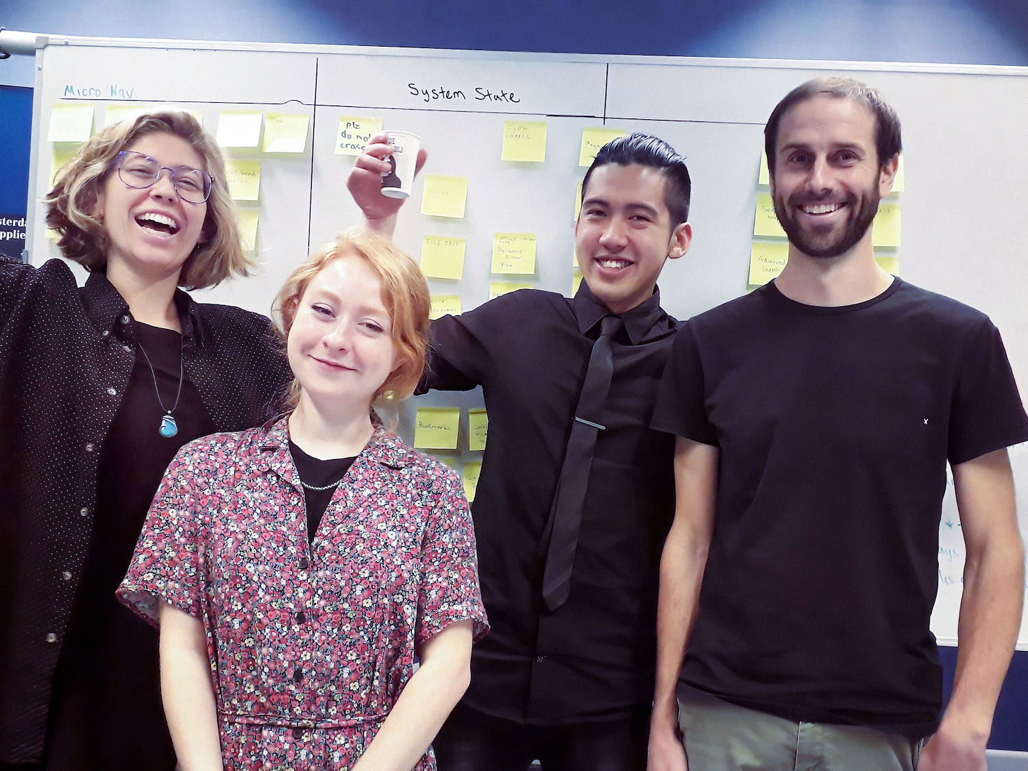 A photo of four members of the Sphere design team (two men and two women) standing in front of a white board with sticky notes on it.